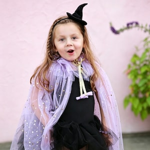 May include: A light purple tulle cape featuring gold star accents, a ruffled collar, and a gold ribbon tie closure. The cape is worn over a black outfit and is paired with a small black witch hat attached to a thin headband with a braided detail.