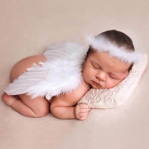 Può includere: Un neonato addormentato, con ali d'angelo di piume bianche e un'aureola abbinata. Il bambino è sdraiato su un cuscino color crema, con uno sfondo morbido e neutro. La pelle del bambino è liscia e l'immagine ha un'atmosfera delicata e serena.