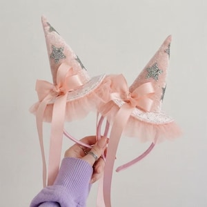 May include: A pair of light pink cone-shaped hats attached to a headband. Each hat features silver glitter star appliques, a ruffled tulle trim at the base, and a long satin ribbon bow in a matching light pink shade that hangs down from the brim.
