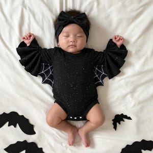 May include: A black long-sleeved bodysuit featuring a subtle white speckled pattern and large, pleated bat-wing sleeves with white spiderweb graphics. The outfit includes a matching black headband with a large bow, worn by an infant.
