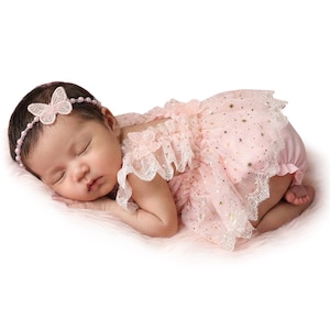 Puede incluir: Un beb&eacute; dormido con un vestido rosa con una capa de tul y detalles de estrellas doradas. El vestido tiene ribete de encaje y aplicaciones de mariposas. Una diadema de mariposa a juego completa el conjunto.