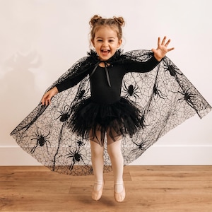 K&ouml;nnte beinhalten: Ein schwarzer Langarm-Body kombiniert mit einem passenden schwarzen T&uuml;ll-Tutu-Rock und einem transparenten schwarzen Umhang mit detailliertem Spinnennetz-Muster und schwarzen Spinnenmotiven. Der Umhang wird am Hals mit einem schwarzen Band gebunden.