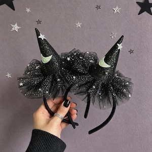 May include: Two black headbands featuring pointed witch hats made of glittery fabric. Each hat is adorned with a pale green crescent moon and a small silver star. The base of each hat is surrounded by layers of black tulle with silver glitter.
