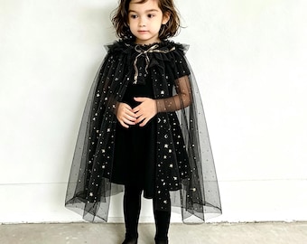 Könnte beinhalten: Ein schwarzer, transparenter Tüllumhang mit goldener Stern- und Mondstickerei, einem gerüschten Kragen und einem metallischen Bandverschluss. Der Umhang wird über einem schlichten schwarzen Kleid und einer schwarzen Strumpfhose getragen.
