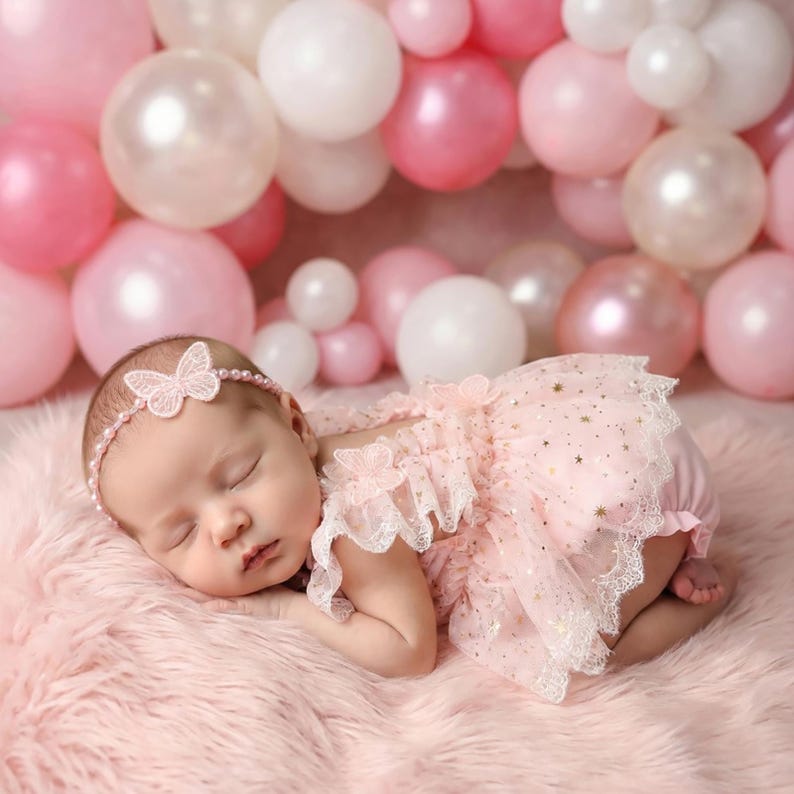 Puede incluir: Un beb&eacute; dormido con un vestido rosa con detalles de estrellas doradas y ribete de encaje, y una diadema de mariposa a juego. El beb&eacute; est&aacute; acostado sobre una superficie rosa y esponjosa, con globos rosas y blancos en el fondo.