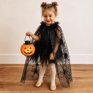 K&ouml;nnte beinhalten: Ein schwarzer T&uuml;llumhang mit Spinnennetz- und Spinnenmuster, getragen &uuml;ber einem schwarzen Trikot mit geschichtetem T&uuml;llrock. Das Outfit wird mit hellen Strumpfhosen und Ballerinas kombiniert, dazu ein leuchtend orangefarbener K&uuml;rbiseimer aus Kunststoff.