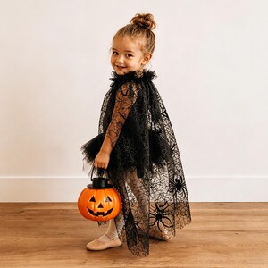 K&ouml;nnte beinhalten: Ein schwarzer, transparenter Umhang mit Spinnennetz- und Spinnenmuster, getragen &uuml;ber einem schwarzen Tutu-Kleid. Das Outfit wird durch einen leuchtend orangefarbenen K&uuml;rbiseimer aus Kunststoff mit einem geschnitzten Jack-o'-Lantern-Gesicht erg&auml;nzt.