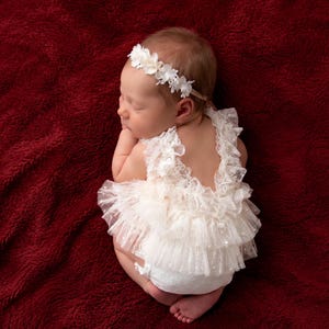 Puede incluir: Un bebé recién nacido durmiendo, vestido con un body de encaje crema con una falda de tul y una diadema floral. El bebé está acurrucado sobre una superficie roja oscura y afelpada. El body presenta un diseño con volantes y delicados detalles de perlas.