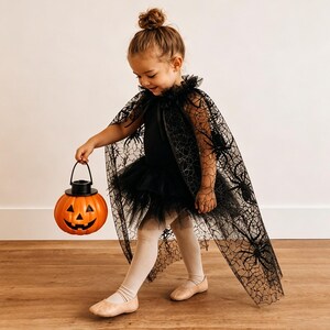 K&ouml;nnte beinhalten: Ein schwarzer, transparenter Umhang mit Spinnennetz- und Spinnenmuster sowie einem R&uuml;schenkragen, getragen &uuml;ber einem schwarzen Trikot mit T&uuml;llrock, kombiniert mit beigen Leggings und Ballerinas, w&auml;hrend ein orangefarbener K&uuml;rbiseimer gehalten wird.