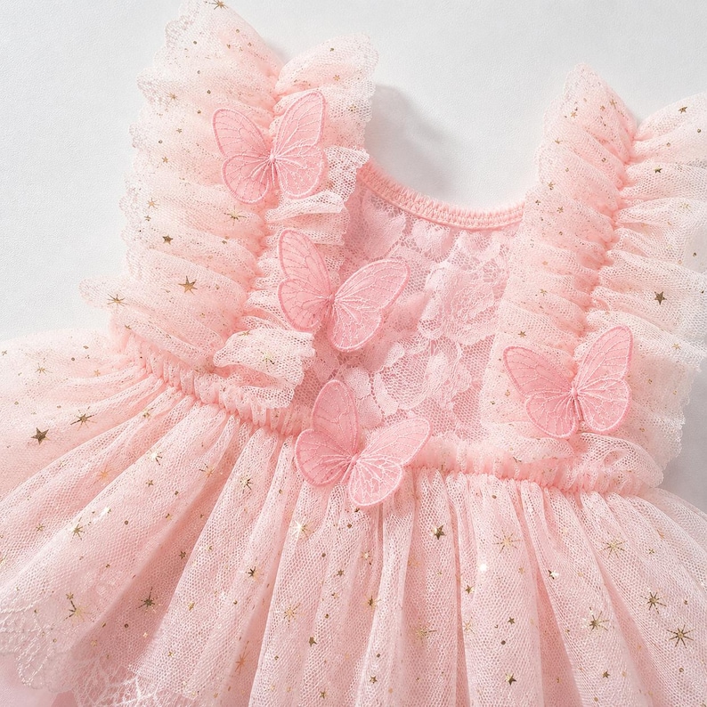 Puede incluir: Vestido rosa claro con falda de tul y corpi&ntilde;o de encaje. El vestido est&aacute; adornado con detalles de estrellas doradas y mariposas rosas bordadas. Tiene mangas con volantes y cuello redondo. El vestido est&aacute; dise&ntilde;ado para una ni&ntilde;a.