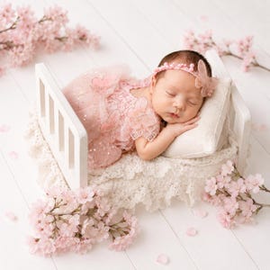 Puede incluir: Un beb&eacute; dormido con un vestido rosa y una diadema con un adorno de mariposa, descansando en una peque&ntilde;a cama blanca con una almohada crema y ropa de cama de encaje. Ramas y p&eacute;talos de flores de cerezo rosas rodean la cama.