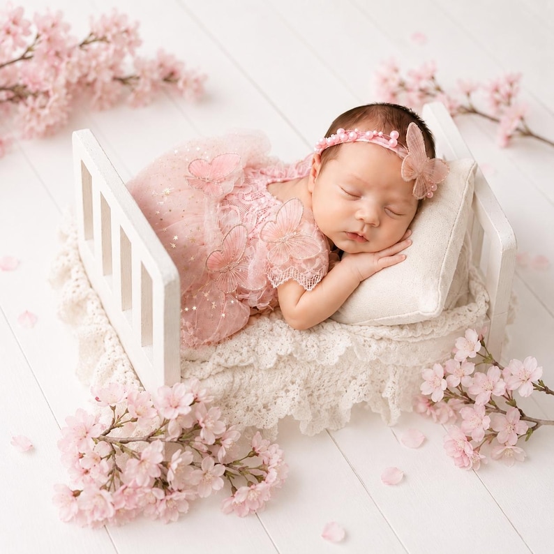 Puede incluir: Un beb&eacute; dormido con un vestido rosa y una diadema con un adorno de mariposa, descansando en una peque&ntilde;a cama blanca con una almohada crema y ropa de cama de encaje. Ramas y p&eacute;talos de flores de cerezo rosas rodean la cama.
