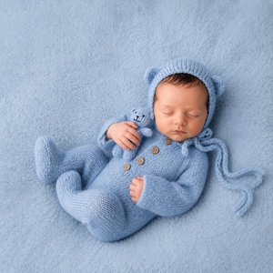 May include: A light blue knitted onesie with long sleeves and feet, featuring four small wooden buttons down the front. The outfit includes a matching bonnet with bear ears and long ties, and a small matching knitted bear toy held by the infant.