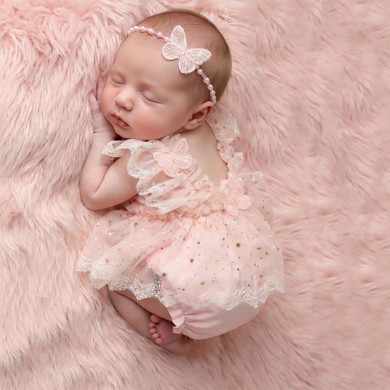 Puede incluir: Un beb&eacute; reci&eacute;n nacido con un pelele rosa con falda de tul y una diadema a juego con dise&ntilde;o de mariposa. El pelele tiene ribete de encaje y detalles de estrellas doradas. El beb&eacute; est&aacute; acostado sobre una superficie rosa esponjosa.