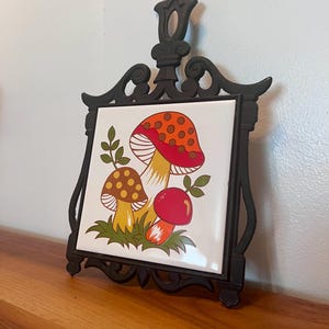 May include: A black metal trivet with a white tile featuring a colorful mushroom design. The mushrooms have red, orange, and brown caps with yellow stems and green leaves. The trivet is on a wooden surface.