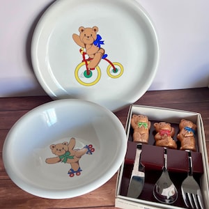 May include: A white children's dinnerware set featuring a plate, bowl, and a boxed set of utensils. The plate has a brown teddy bear riding a bicycle. The bowl has a teddy bear on roller skates. The utensils have teddy bear toppers.