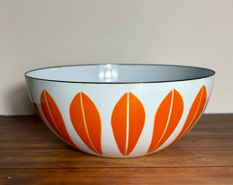 May include: A large, round, white enamel bowl featuring a repeating pattern of vibrant orange, leaf-shaped motifs around the exterior. The bowl has a thin, dark rim along the top edge, providing a clear contrast against the white interior and exterior.