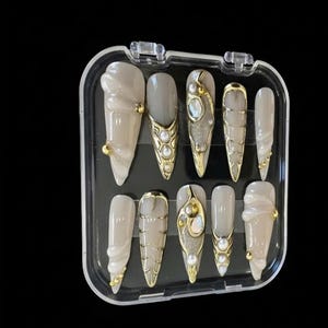May include: A set of ten artificial nails in a clear plastic case. The nails are various shades of beige and gray, with gold and pearl accents. Some nails have a textured design.