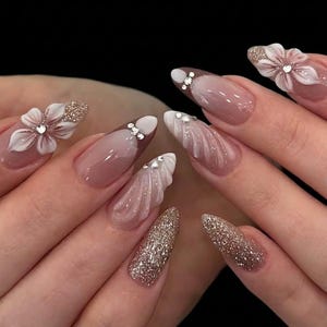 May include: Elegant, long, almond-shaped artificial nails. Some nails feature a glittery gold finish, while others showcase a soft pink base with white tips and delicate floral embellishments. Rhinestones add sparkle to the design.