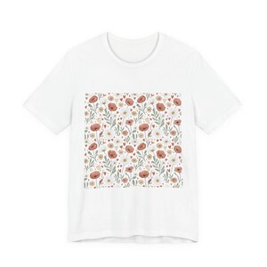 Könnte beinhalten: Weißes T-Shirt mit einem floralen Muster aus roten Mohnblumen, weißen Gänseblümchen und kleinen gelben Herzen. Das Design umfasst grüne Blätter und Stiele und erzeugt einen skurrilen, von der Natur inspirierten Druck. Das Shirt besteht aus einem weichen, bequemen Material.