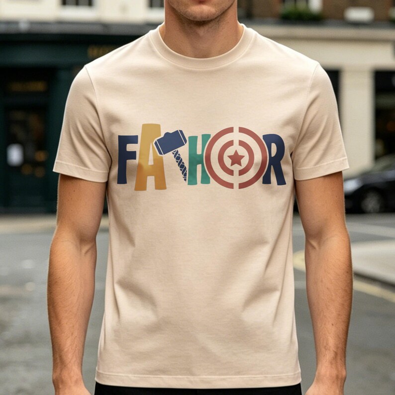 May include: Beige short-sleeve t-shirt with the word "FATHOR" in colorful block letters. The "A" is a hammer, the "H" is a lightning bolt, and the "O" is a target with a star in the center. The shirt is made of a soft material.
