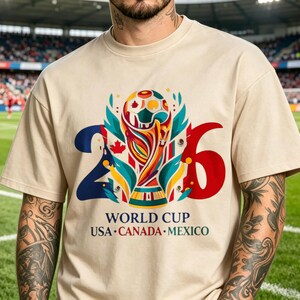 Puede incluir: Una camiseta beige de cuello redondo con un gr&aacute;fico del trofeo de la Copa Mundial 2026 centrado entre los n&uacute;meros 2 y 6. El dise&ntilde;o incluye formas abstractas coloridas, hojas de arce y el texto "WORLD CUP USA CANADA MEXICO" debajo.