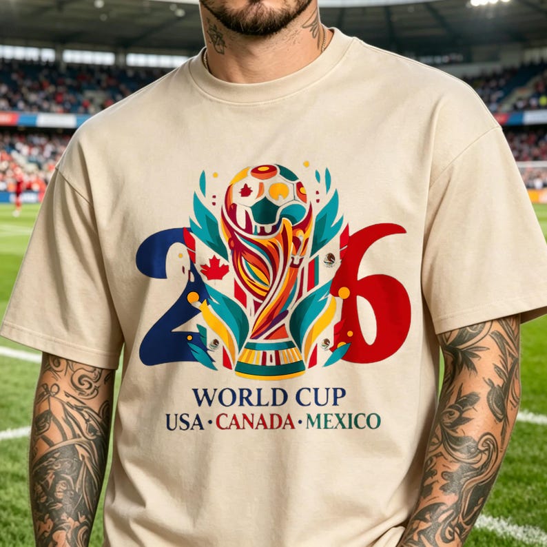 Puede incluir: Una camiseta beige de cuello redondo con un gr&aacute;fico del trofeo de la Copa Mundial 2026 centrado entre los n&uacute;meros 2 y 6. El dise&ntilde;o incluye formas abstractas coloridas, hojas de arce y el texto "WORLD CUP USA CANADA MEXICO" debajo.