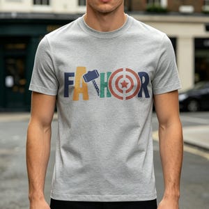 May include: A gray t-shirt with the word "FA THOR" printed across the chest in colorful block letters. The letters are designed with superhero-themed graphics, including a hammer, a target, and a saw.