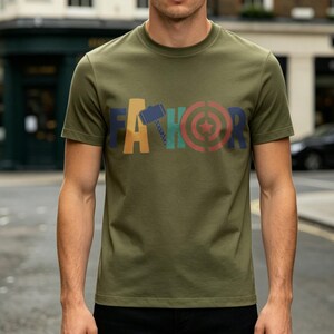 May include: Olive green t-shirt with the word "FATHOR" in colorful block letters. The "A" is yellow, the "H" is blue, the "O" is red, and the "R" is a mix of red and green. The shirt has short sleeves and a crew neck.