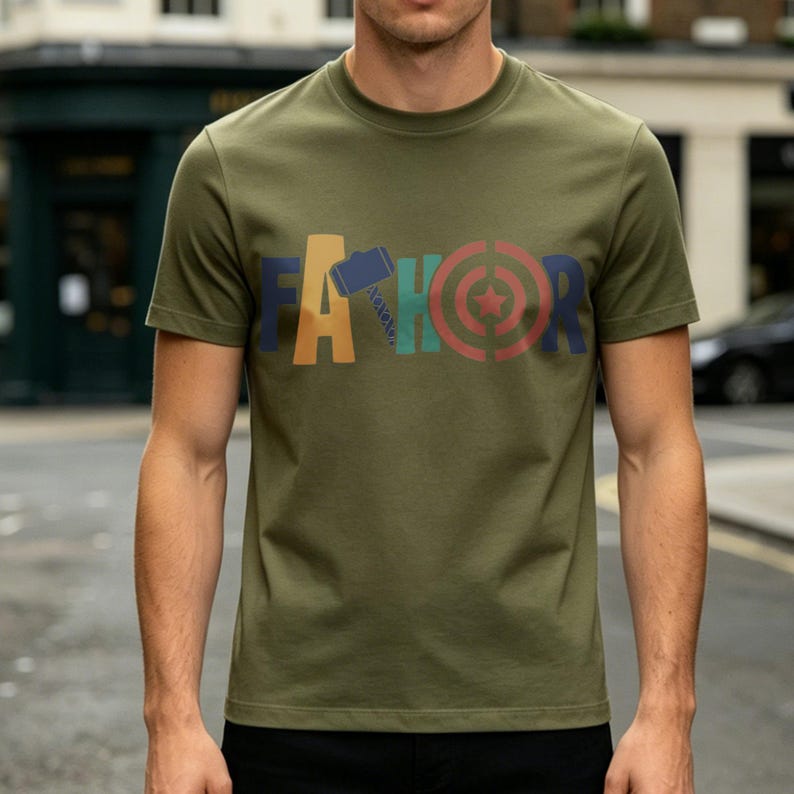 May include: Olive green t-shirt with the word "FATHOR" in colorful block letters. The "A" is yellow, the "H" is blue, the "O" is red, and the "R" is a mix of red and green. The shirt has short sleeves and a crew neck.