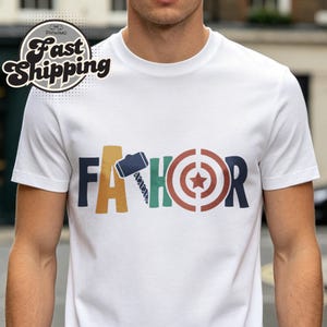 May include: White t-shirt with the word "FATHOR" in colorful block letters. The "A" is a hammer, the "H" is a green and blue design, and the "O" is a target with a star in the center.
