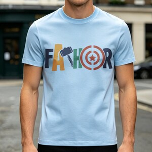 May include: Light blue t-shirt with the word "FATHOR" in colorful letters. The "A" is a hammer, the "H" is a saw, and the "O" is a target with a star. The shirt is short-sleeved and made of a soft material.
