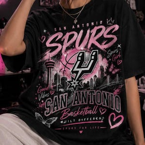May include: A black short-sleeve t-shirt featuring a graphic print with the text San Antonio Spurs, San Antonio Basketball, Built Different, and Spurs For Life. The design includes pink hearts, butterflies, a city skyline, and the Spurs logo.