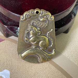 May include: A brass-colored pendant featuring a detailed relief of a rat, perched on a stylized branch. The pendant has an ornate border with a small hole at the top for hanging. The background is a gradient of brown and gold.