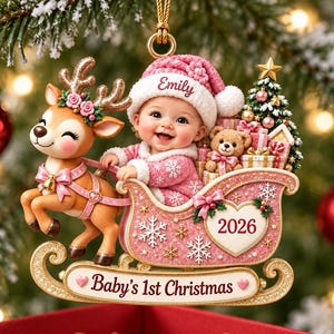 May include: A Christmas ornament featuring a baby in a pink hat with the name Emily, sitting in a pink sleigh pulled by a reindeer. The sleigh is decorated with snowflakes, a heart labeled 2026, and a banner reading Baby's 1st Christmas.