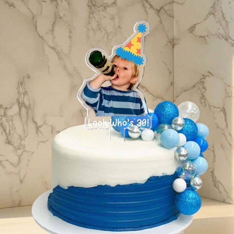 May include: A birthday cake with white frosting and a blue bottom layer. The cake is decorated with blue and silver ball ornaments. A clear acrylic cake topper features a photo of a person drinking from a bottle, wearing a party hat, and the text "Look Who's 30!"