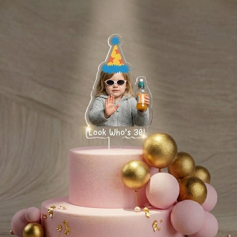 May include: A pink two-tiered birthday cake with gold and pink decorative spheres. A clear acrylic cake topper features a photo of a child wearing sunglasses and a party hat, holding a bottle, with the text "Look Who's 30!"
