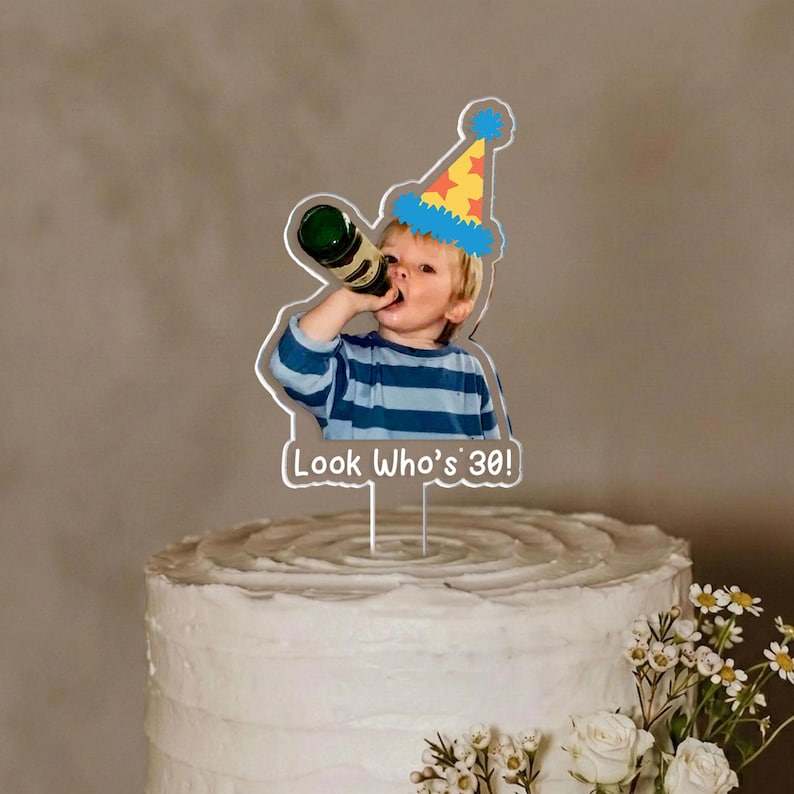 May include: A cake topper featuring a cartoon image of a child drinking from a bottle, wearing a party hat. The text "Look Who's 30!" is below the image. The topper is placed on a white frosted cake with white flowers.