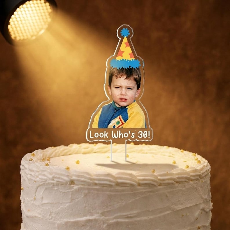 May include: A birthday cake with white frosting and gold sprinkles, topped with a clear acrylic cake topper. The topper features a photo of a person wearing a party hat and the text "Look Who's 30!"