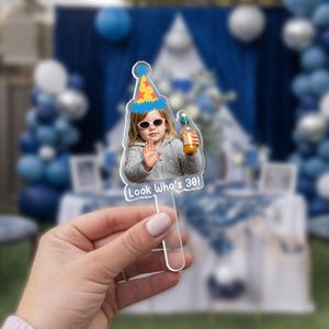 May include: Clear acrylic cake topper featuring a photo of a person wearing sunglasses and a party hat, holding a bottle. The text "Look Who's 30!" is printed on the bottom. The background is a blurred outdoor party scene.