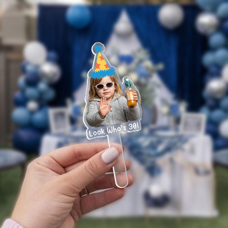 May include: Clear acrylic cake topper featuring a photo of a person wearing sunglasses and a party hat, holding a bottle. The text "Look Who's 30!" is printed on the bottom. The background is a blurred outdoor party scene.