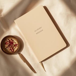 May include: A beige notebook with the words "ALLRADY ENOUGH" printed on the cover, a gold pen, and a small gold dish with dried flowers. The items are arranged on a cream-coloured surface.
