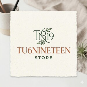 May include: A square, off-white card with the text "TN19" and "TU6NINETEEN STORE" in a mix of green and brown fonts. The logo includes a leaf design. The card is on a white surface with a neutral-colored cloth and a small plant.