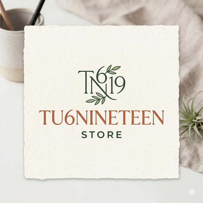 May include: A square, off-white card with the text "TN19" and "TU6NINETEEN STORE" in a mix of green and brown fonts. The logo includes a leaf design. The card is on a white surface with a neutral-colored cloth and a small plant.