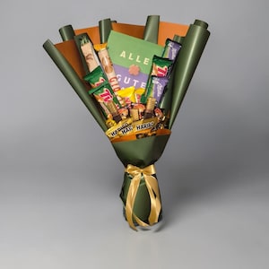 Könnte beinhalten: Ein Süßigkeitenstrauß mit verschiedenen Schokoriegeln und Süßigkeiten, verpackt in grünem und orangefarbenem Papier und mit einer goldenen Schleife gebunden. Der Strauß enthält Haribo und Schokoriegel, mit einer grünen Karte mit der Aufschrift "ALLE GUTE".
