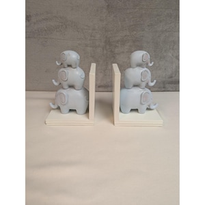 May include: A pair of decorative bookends featuring a stack of three light blue elephants. The elephants have pink accents and are perched on a white base. The bookends are designed to hold books upright and add a whimsical touch to any room.