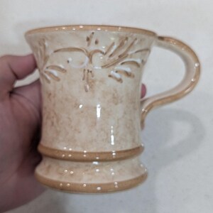 May include: A beige ceramic mug with a handle. The mug features a decorative embossed floral design and a brown band around the base. The rim and handle are also brown. The mug is held by a hand.