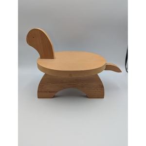 May include: A light brown wooden turtle-shaped stand featuring a smooth circular top, a curved base with an arched cutout, a small head with a single dot for an eye, and a pointed tail. The natural wood grain is visible across the entire structure.