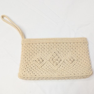 May include: A rectangular beige macrame clutch bag featuring a wrist strap and a repeating diamond pattern with central knot clusters. The accessory is crafted from woven cord, showcasing a textured, open-work design across the entire front surface.