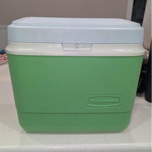 May include: A rectangular Rubbermaid cooler featuring a vibrant green body with a textured finish and a light blue lid. The front of the cooler displays the embossed Rubbermaid brand name in a rectangular recessed panel, with a white trim border.